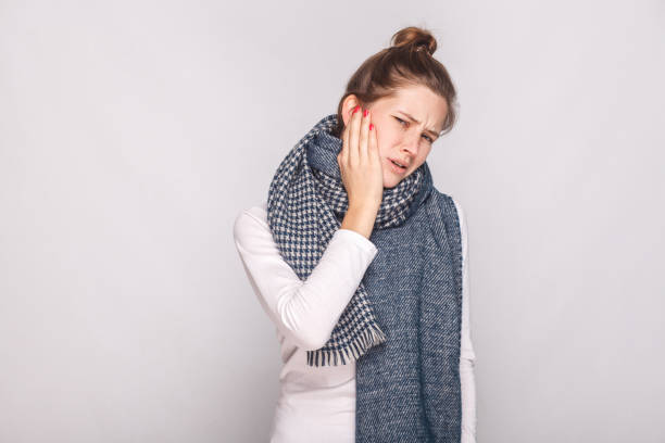 A woman wearing a long blue, gray, and white scarf, grasps her jaw in pain.